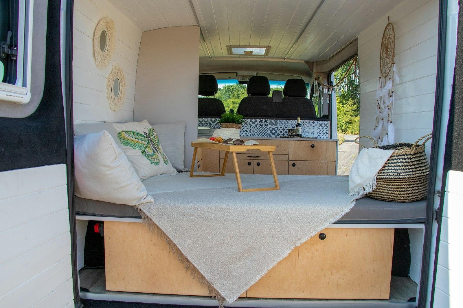 Converted campervan interior with an upholstered bed platform, cushions and a small table, cab seats in the background