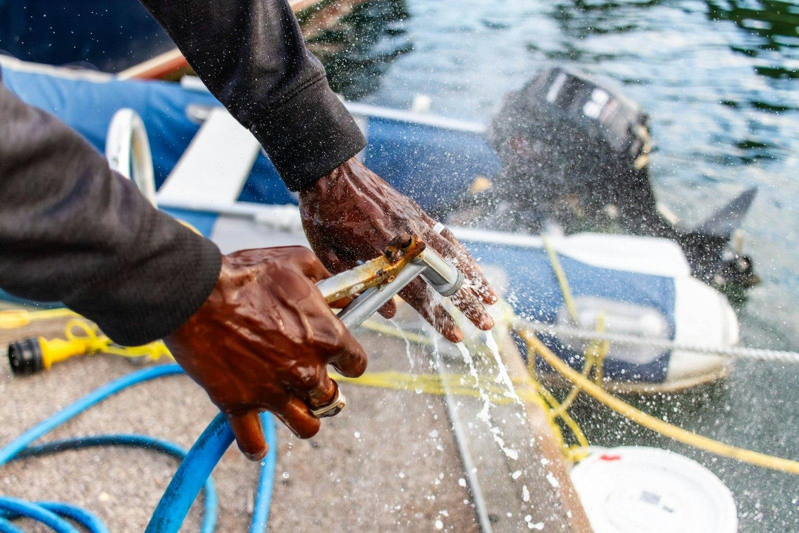 Hands holding a hose nozzle spraying fresh water near a small boat at a dock