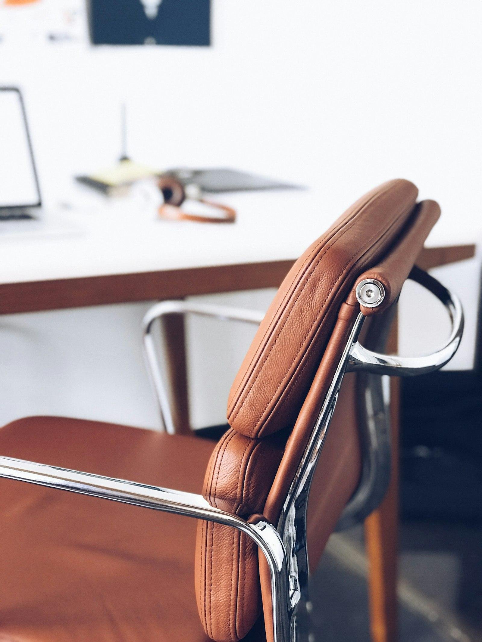Close-up of a tan leather office chair with chrome arms beside a white desk