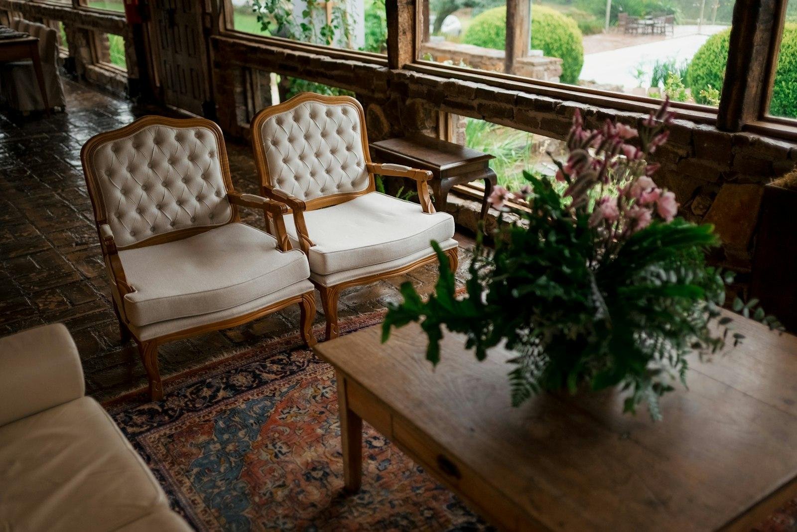 Two cream button-tufted armchairs with carved timber frames beside a coffee table in a sunlit room